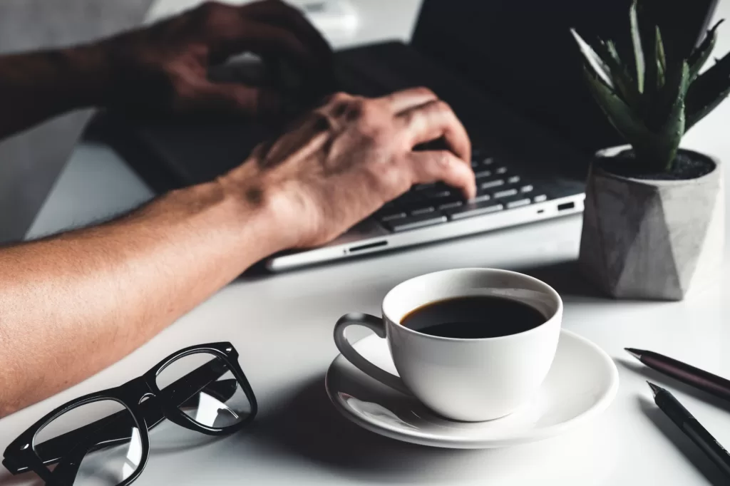 A man types on a laptop, business concept, glasses, a cup of coffee and a pen on a gray background.