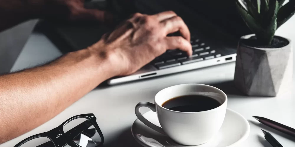 A man types on a laptop, business concept, glasses, a cup of coffee and a pen on a gray background.